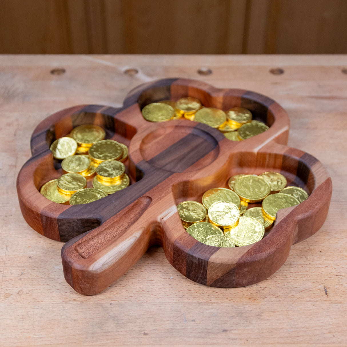 Shamrock-shaped wooden tray holding gold foil chocolate coins on a workbench.