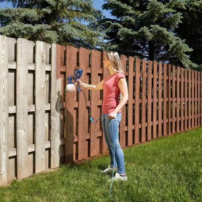 Woman using paint sprayer to stain wooden fence outdoors, demonstrating fence painting tool.