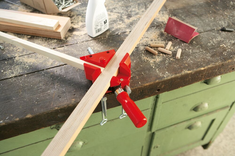 Woodworking clamp holding two pieces of wood on a workbench with tools in the background.