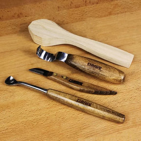 Three wood carving tools with wooden handles and a carved wooden spoon blank on a wood surface
