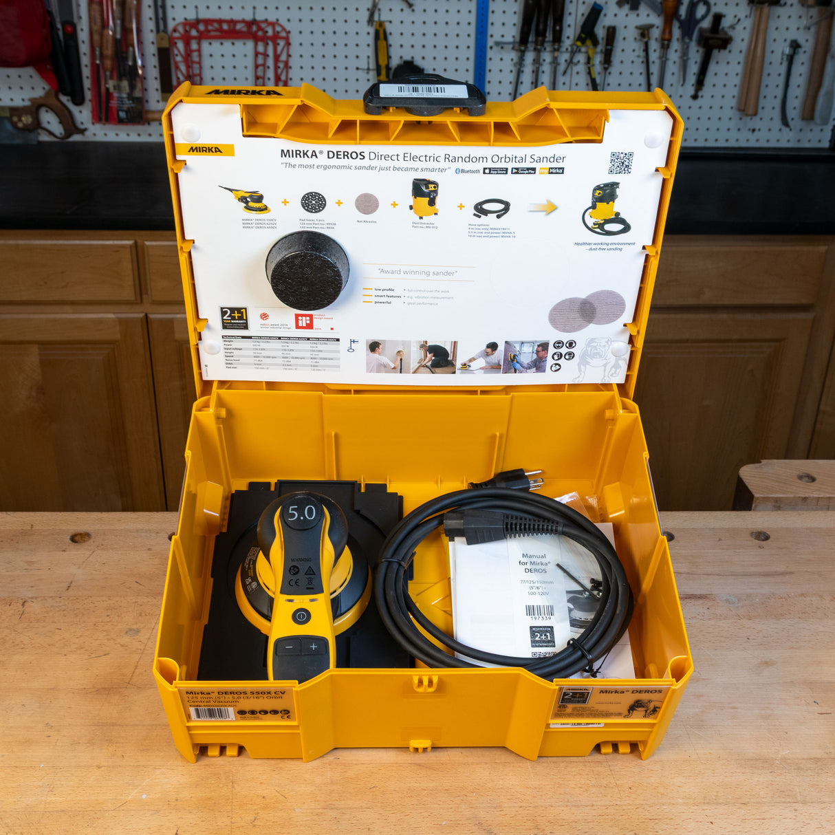 Mirka electric random orbital sander in a yellow case on a wooden table with a workshop background.