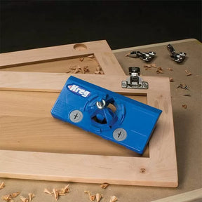 Blue concealed hinge jig tool on unfinished cabinet door with hardware and wood shavings