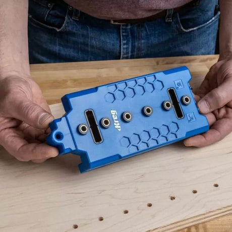Hands holding a blue pocket hole jig on a wooden workbench for precise woodworking joints
