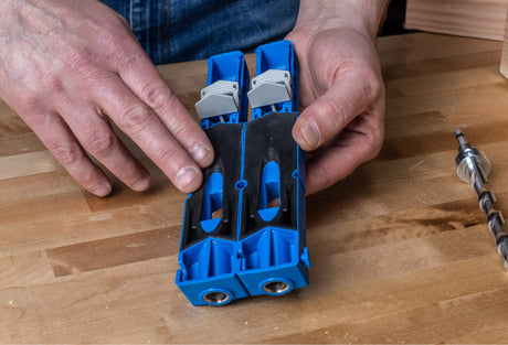 Person holding a blue pocket hole jig on a woodworking bench with drill bit nearby