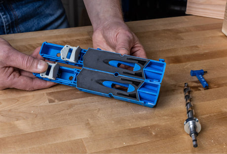Hand holding a blue pocket hole jig with a drill bit and depth collar on a woodworking bench.