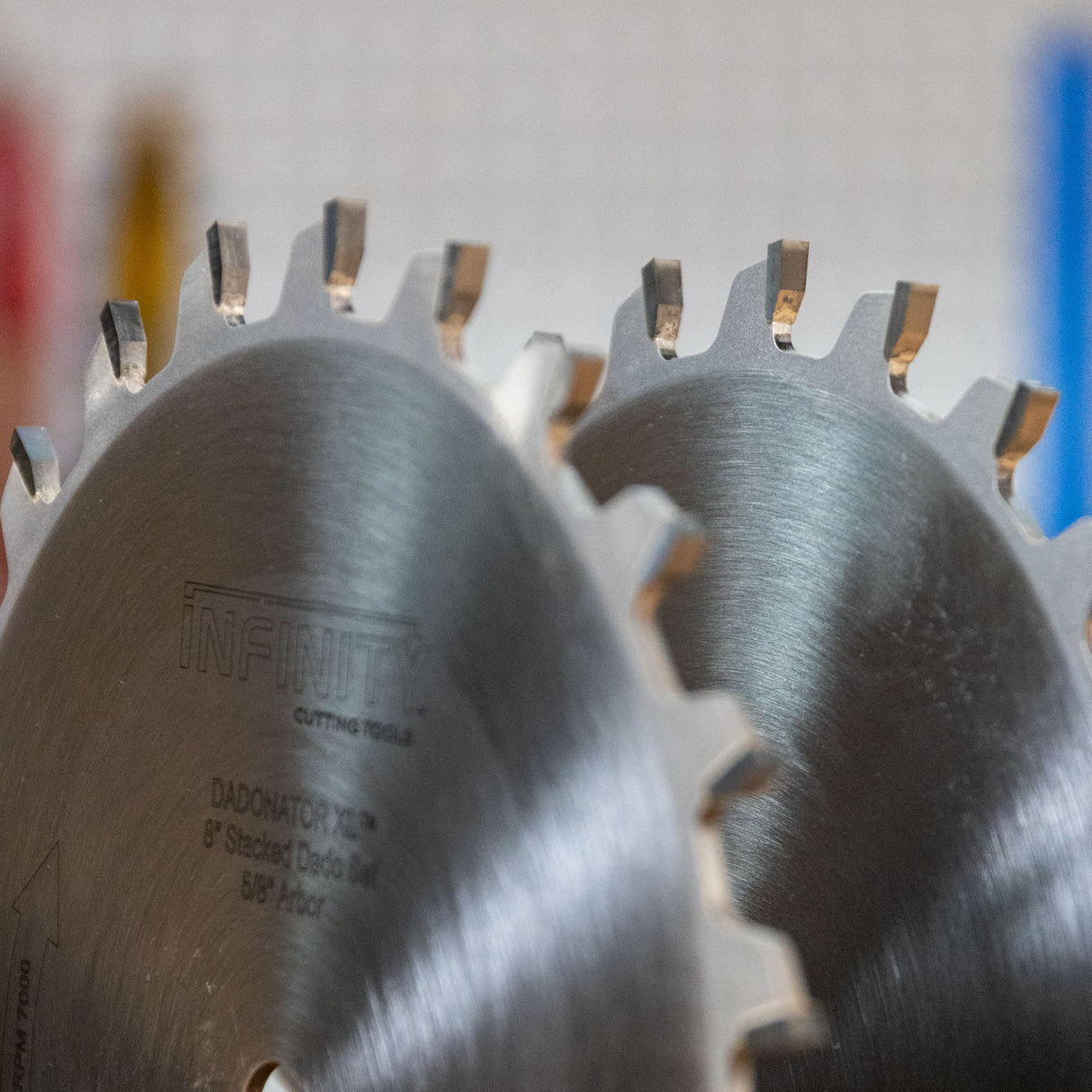 Two Infinity brand circular saw blades with stacked diamond edges, shown in a close-up shot with a blurred workshop background.