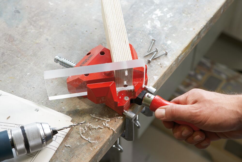 Person using a red Bessey corner clamp on a workbench with a drill on bench.